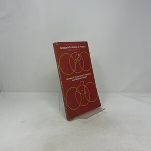 May include: A red book titled "Graduate Problems in Physics With Solutions" by Jeremiah A. Cronin, David F. Greenberg, and Valentine L. Telegdi. The cover features overlapping circles in pink and green. The book is propped up on a clear plastic stand.