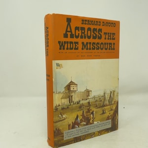May include: A hardcover book titled Across the Wide Missouri by Bernard DeVoto with an orange spine and cover. The cover features a historical painting of a fort, teepees, and figures on horseback, with text detailing the Miller collection.
