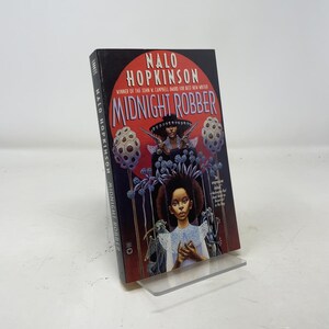 May include: A hardcover book titled "Midnight Robber" by Nalo Hopkinson. The cover features a woman with a hat and guns, and a young girl. The book is displayed on a clear acrylic stand.