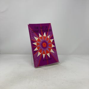 May include: A hardcover book titled "THE ART OF CHART SYNTHESIS" with a purple cover. The cover features a geometric design in orange, white, and red. The book is propped up on a clear stand.