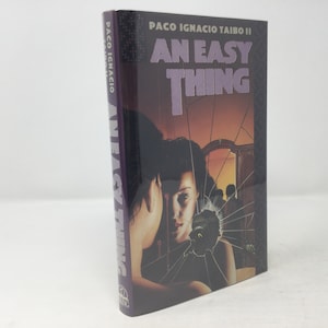 May include: A purple hardback book titled "An Easy Thing" by Paco Ignacio Taibo II. The cover features a woman looking into a broken mirror with a dark background.