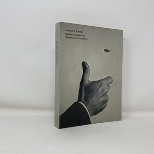 May include: A hardcover book titled "Statistical Analysis for Business and Economics" by Leonard J. Kazmier. The cover features a black and white image of a hand giving a thumbs-up gesture, with a coin in the air. The book is on a white surface.