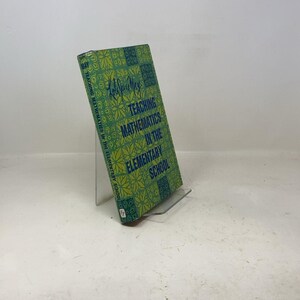 May include: A vintage book titled "Teaching Mathematics in the Elementary School" is displayed on a clear stand. The book has a green cover with a floral pattern and blue text. The author's signature is visible.