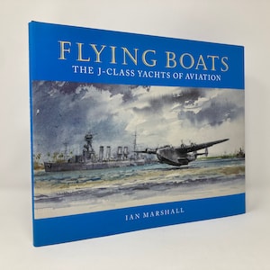 Pode incluir: Um livro de capa dura intitulado "FLYING BOATS The J-Class Yachts of Aviation" de Ian Marshall. A capa apresenta uma pintura em aquarela de um hidroavião e um navio na água, contra um céu nublado. O livro tem uma capa azul.