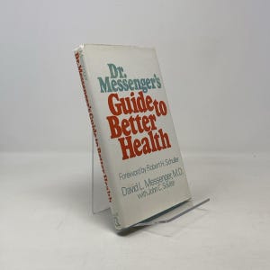 May include: A book titled "Dr. Messenger's Guide to Better Health" is displayed on a clear acrylic stand. The book's cover is white with blue and red text. The book is angled slightly, showcasing the title and author. The book is in cm.