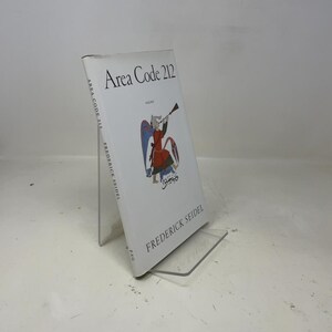 Puede incluir: Un libro de tapa dura titulado "Area Code 212" de Frederick Seidel. La portada es blanca con texto negro y una ilustración colorida de una figura tocando una trompeta. El libro está apoyado en un soporte de plástico transparente.