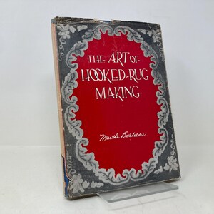 May include: A vintage book titled "The Art of Hooked-Rug Making" by Martha Babbitt. The cover features a decorative floral design in gray and white against a red background.