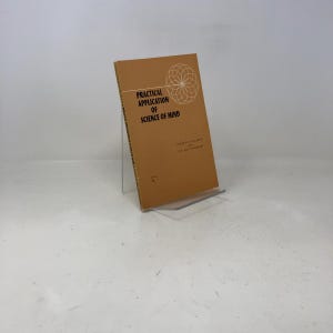 以下が含まれることがあります： 「Practical Application of Science of Mind」というタイトルの本が、透明なアクリルスタンドに展示されています。本は、白い文字と幾何学模様が施された茶色の表紙です。本はわずかに傾いています。