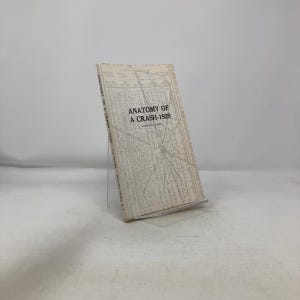 May include: A hardcover book titled "Anatomy of a Crash-1929" is displayed on a clear stand. The book's cover features a light-colored background with text and a graphic resembling a shattered window. The book is by L.A. Levin.