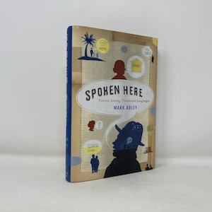 May include: A hardcover book titled "Spoken Here" with a blue spine. The cover features illustrations of silhouettes, speech bubbles, and a palm tree. The book's title is in a white oval, with the author's name below.