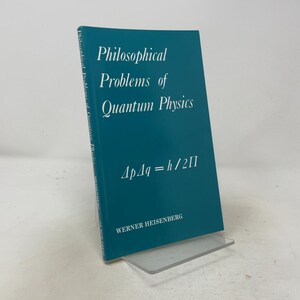 May include: A teal softcover book titled Philosophical Problems of Quantum Physics by Werner Heisenberg. The cover features white text and the mathematical equation ΔpΔq = h / 2Π centered on the front. The spine also displays the title.