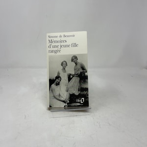 May include: A paperback book titled "Mémoires d'une jeune fille rangée" by Simone de Beauvoir. The cover features a black and white photograph of three women in vintage dresses. The book is displayed on a clear plastic stand.