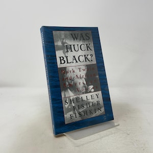 Op de afbeelding: Een hardcover boek getiteld Was Huck Black? Mark Twain and African American Voices door Shelley Fisher Fishkin. De kaft heeft een blauwe textuurrand en een centrale zwart-witfoto van een persoon, met dikgedrukte zwarte tekst.