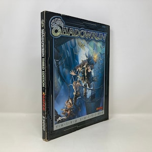 Puede incluir: Un libro negro con una cubierta plateada y azul que presenta un dragón y el texto "Shadowrun Tercera Edición" y "KanPR-25000".