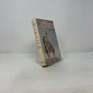 May include: A vintage book titled "The Hands of Cantii" by Tom Lea. The cover features an illustration of a person on horseback. The book has a light-colored spine with text and a decorative emblem. The book is propped up on a clear stand.