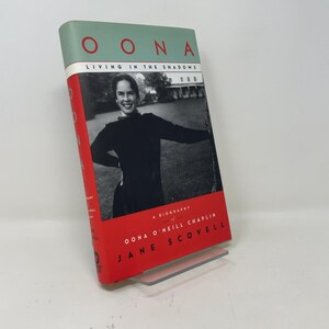 May include: A hardcover book titled Oona: Living in the Shadows, a biography of Oona O'Neill Chaplin by Jane Scovell. The cover features a black and white photograph of a smiling person, with a sage green top section and a bright red border.