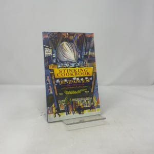 Puede incluir: Un libro de cocina titulado "The Stinking Cookbook" de The Stinking Rose. La portada presenta una ilustración de un gran bulbo de ajo sobre un fondo de paisaje urbano. El libro se exhibe en un soporte de plástico transparente.