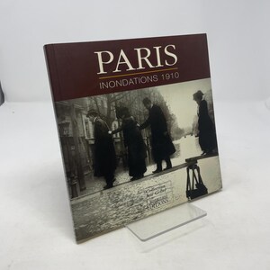 May include: A hardcover book titled "PARIS INONDATIONS 1910" with a sepia-toned photograph of people walking on a makeshift bridge. The book has a maroon cover and is displayed on a clear acrylic stand. The book's title is in large white letters.