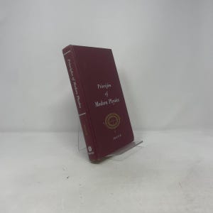 Puede incluir: Un libro granate titulado "Principles of Modern Physics" con letras doradas y un gráfico en forma de brújula. El libro está apoyado en un soporte de plástico transparente, mostrando su portada y lomo. El libro es publicado por Wiley.
