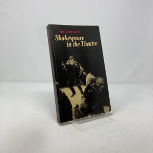 May include: A hardcover book titled "Shakespeare in the Theatre" by Richard David. The cover is black with a sepia-toned illustration of a man holding a sword. The book is propped up on a clear plastic stand.