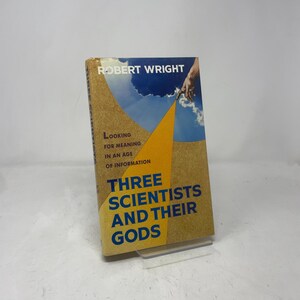 Op de afbeelding: Een boek van Robert Wright, getiteld "Three Scientists and Their Gods". De omslag heeft een gele driehoek op een bruine achtergrond, met blauwe tekst en een luchtillustratie.