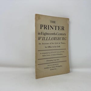 May include: A vintage book titled "THE PRINTER in Eighteenth-Century WILLIAMSBURG." The cover is a faded tan color with black text. The book details the life, times, office, and craft of a printer in Williamsburg.