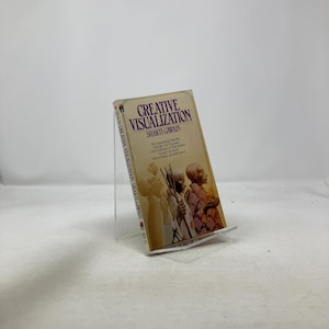 May include: A paperback book titled "Creative Visualization" by Shakti Gawain. The cover has a beige background with the title in purple and the author's name in black. The book is displayed on a clear acrylic stand.