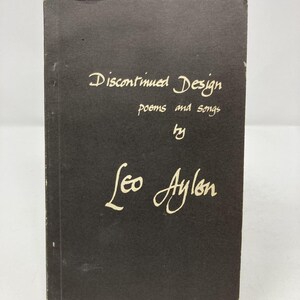 May include: A black book titled "Discontinued Design" with the subtitle "poems and songs" by Leo Aylon. The text is in a handwritten style and is in a cream color. The book is standing upright.