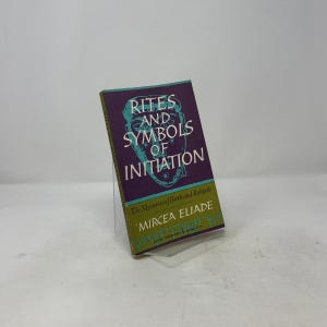 May include: A paperback book titled Rites and Symbols of Initiation by Mircea Eliade. The cover features a purple background with a teal line-art face illustration, white text, and a green lower section with the subtitle The Mysteries of Birth and Rebirth.