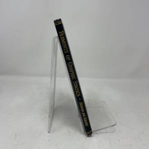Può includere: Un dorso di libro nero intitolato Elements of Graphic Statics di Hudson e Squire, con scritte dorate. Il libro è posizionato verticalmente su un supporto in acrilico trasparente, mostrando il bordo laterale con sottili linee dorate orizzontali.