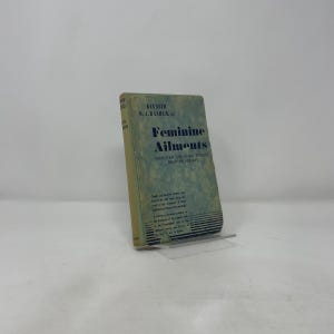 May include: A vintage book titled "Feminine Ailments" by Kenneth D.A. Bashem. The book has a light green cover with blue text and is propped up on a clear stand. The book's title is prominently displayed.