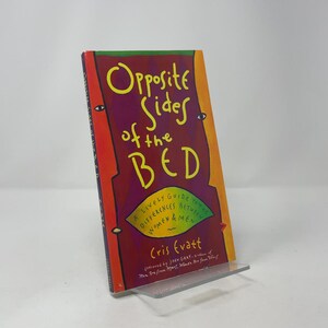 以下が含まれることがあります： クリス・エヴァット著の「Opposite Sides of the Bed」というタイトルの本。表紙は赤、紫、オレンジのミックスで、タイトルは黄色です。本は透明なプラスチック製のスタンドに立てかけられています。副題は「A Lively Guide to the Differences Between Women & Men」です。