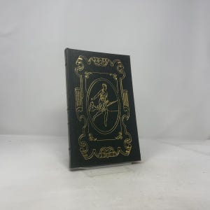 May include: A dark green book with gold embossed detailing. The cover features a gold framed oval with a figure holding a flowing object. The book has a textured appearance and is standing upright.