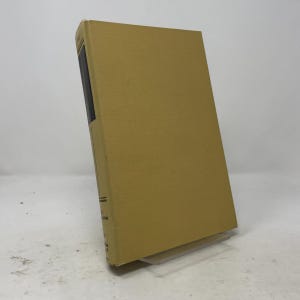 Op de afbeelding: Een hardcover boek met een effen beige stoffen kaft. De rug van het boek is zichtbaar, met een donker rechthoekig paneel met zwarte tekst en horizontale lijnen. Het boek staat rechtop op een doorzichtige plastic standaard voor weergave.