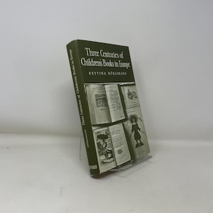 Pode incluir: Um livro de capa dura intitulado "Three Centuries of Children's Books in Europe" de Bettina Hürlimann. O livro tem uma capa verde escura com texto e ilustrações brancas. O livro é exibido em um suporte transparente.