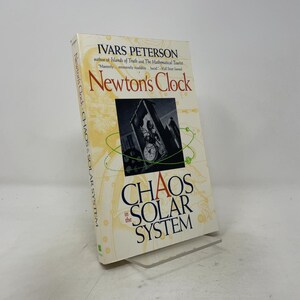May include: A hardcover book titled Newton's Clock: Chaos in the Solar System by Ivars Peterson. The cover features a cream background with faint gold celestial illustrations, a central black and white image of a clock, and green orbital lines.