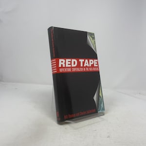 May include: A hardcover book titled "RED TAPE" with a black cover and red lettering. The book's subtitle reads "Adventure Capitalism in the New Russia." The author's names, Bill Thomas and Charles Sutherland, are printed at the bottom.