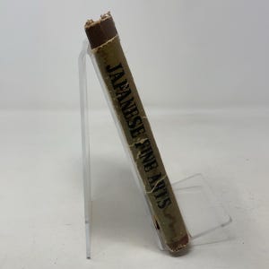 May include: A vintage book titled "Japanese Fine Arts" is displayed on a clear acrylic stand. The book has a worn, tan cover with the title in black lettering. The spine is dark brown and shows signs of age.