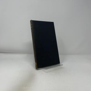May include: A hardcover book with a black cover and gold lettering on the spine. The book is propped up on a clear plastic stand against a white background. The book's edges are a dark brown color.