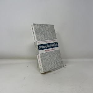 Puede incluir: Un libro de tapa dura titulado "Breaking the Maya Code" de Michael D. Coe. El libro tiene una cubierta blanca con un patrón gris y el título en azul y rojo. El libro está apoyado en un soporte transparente.
