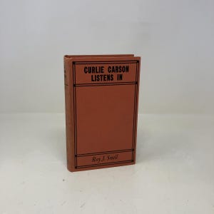 Puede incluir: Un libro antiguo titulado "Curlie Carson Listens In" de Roy J. Snell. El libro tiene una cubierta naranja sólida con texto negro y detalles de borde. El libro está de pie sobre un fondo blanco liso.