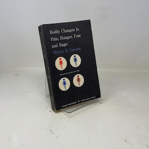 Puede incluir: Un libro de tapa dura titulado "Bodily Changes In Pain, Hunger, Fear and Rage" de Walter B. Cannon. El libro tiene una cubierta negra con texto blanco y cuatro ilustraciones circulares de figuras de palo en rojo y azul. El libro está apoyado en un soporte de plástico transparente.