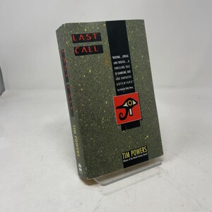 May include: A hardcover book titled Last Call by Tim Powers with a dark green speckled cover. The front features a black vertical stripe, a red square with an Eye of Horus symbol, and text that reads Riveting... Lyrical and Brutal.