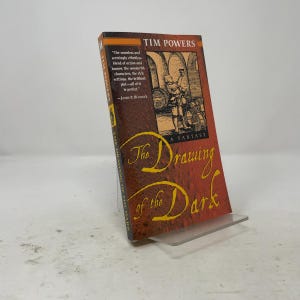 May include: A paperback book titled The Drawing of the Dark by Tim Powers. The cover features a red and brown textured background with a black-and-white illustration of a person holding a mug in a cellar. The title is written in yellow script.