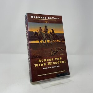 Könnte beinhalten: Ein gebundenes Buch mit dem Titel "Across the Wide Missouri" von Bernard DeVoto. Das Cover zeigt ein Gemälde von Reitern, mit dem Titel und dem Namen des Autors. Das Buch hat ein Mariner Books-Logo.