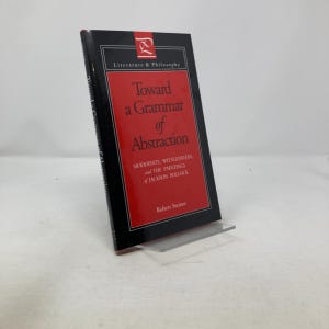 以下が含まれることがあります： ロバート・シュタイナー著「Toward a Grammar of Abstraction」というタイトルの本。本は、白いタイトルが入った赤い長方形の黒い表紙です。本は透明なプラスチック製のスタンドに立てかけられています。
