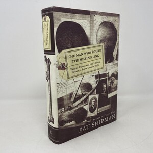 May include: A hardcover book titled "The Man Who Found The Missing Link" by Pat Shipman. The cover features sepia-toned images and text, including a portrait and illustrations. The book's spine is visible.