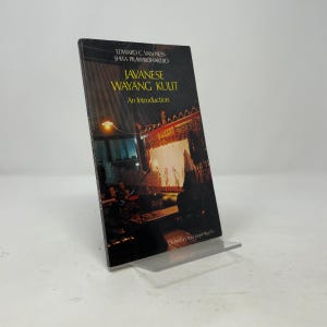 Puede incluir: Un libro de bolsillo titulado Javanese Wayang Kulit: An Introduction por Edward C. Van Ness y Shita Prawirohardjo. La portada presenta una fotografía oscura y atmosférica de una representación tradicional de teatro de sombras con luz cálida.