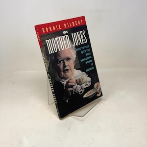 Puede incluir: Se muestra un libro titulado "Ronnie Gilbert sobre Mother Jones". La portada presenta un retrato de una mujer mayor sosteniendo una taza de té. El lomo del libro es rojo y el título está en letras blancas grandes.