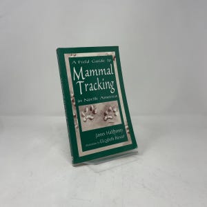 Può includere: Una guida sul campo verde intitolata "A Field Guide to Mammal Tracking in North America". La copertina presenta il titolo in bianco, un'illustrazione di impronte di zampe e informazioni sull'autore. Il libro è leggermente inclinato a sinistra.